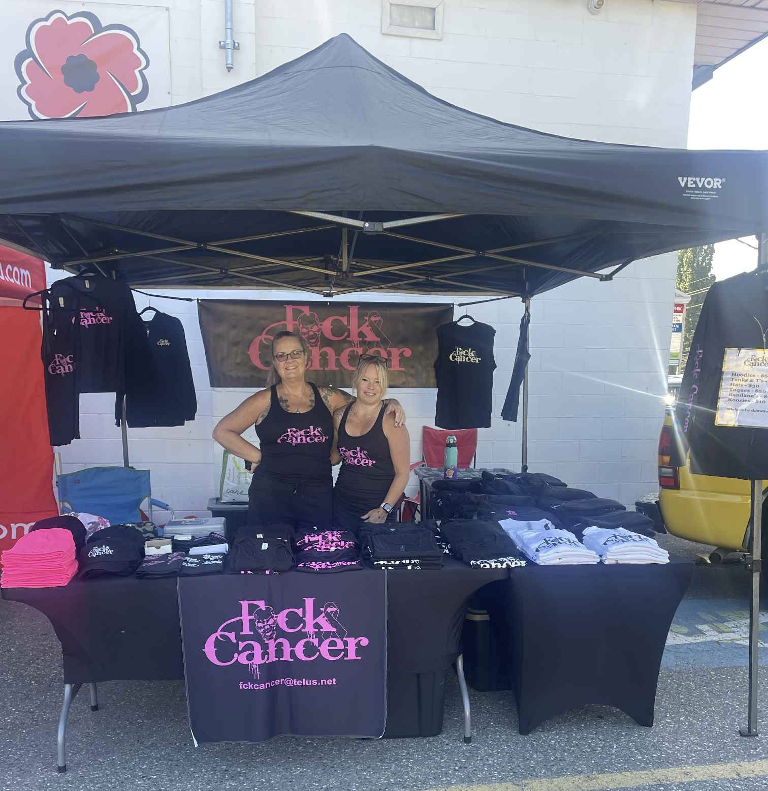 Two volunteers at an FCK Cancer merch table under a tent