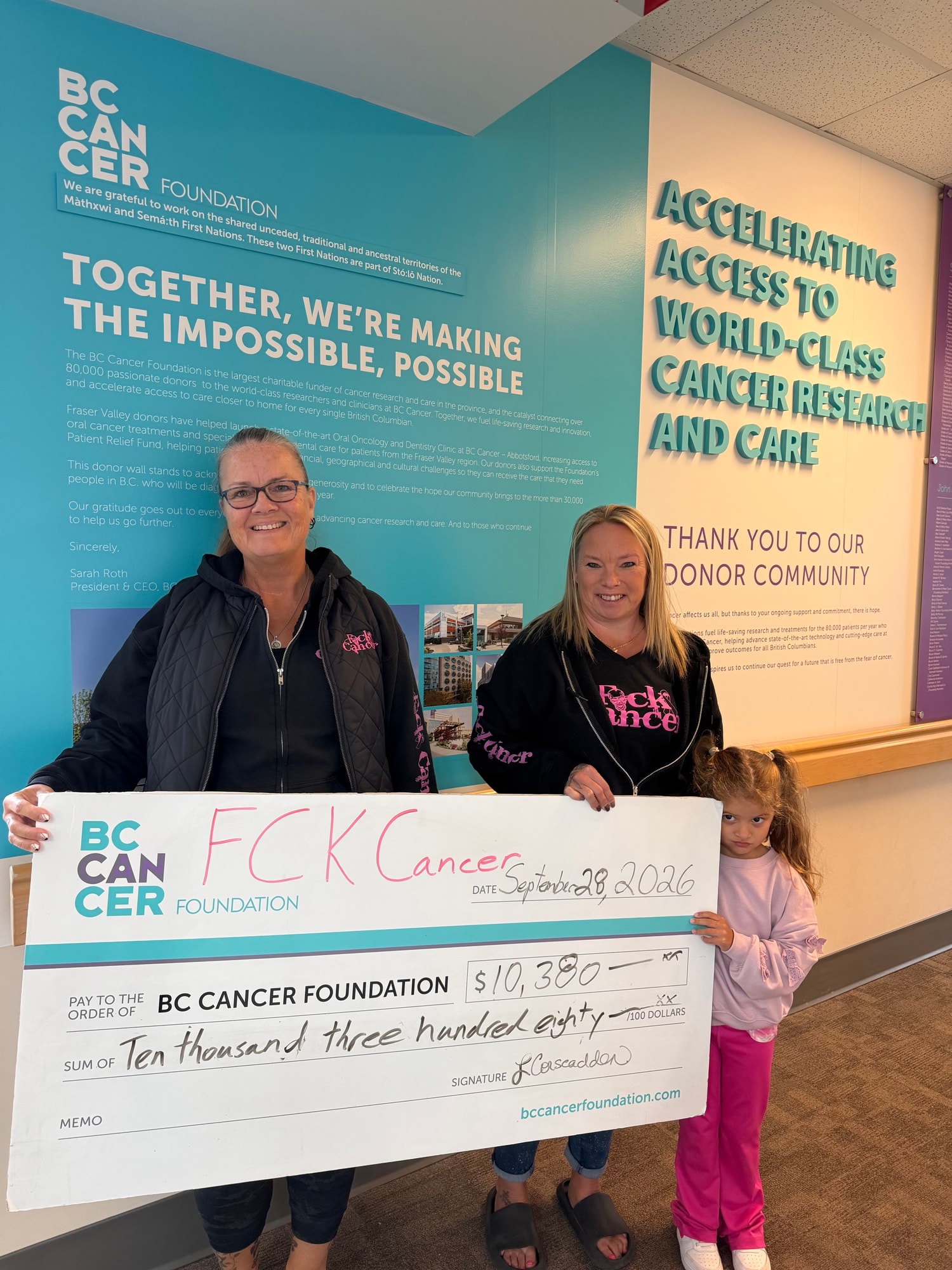 Two women and a girl holding a $10,380 cheque at a BC Cancer Foundation wall
